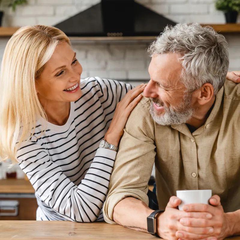 An older couple smiling at each other while in a kitchen.