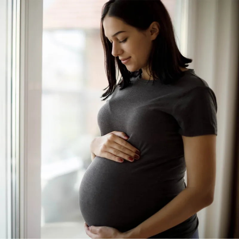 A pregnant young woman holds her belly standing near a window