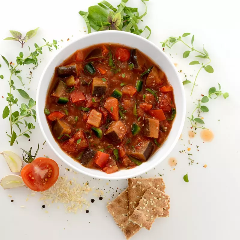 Bowl of vegetable ragout surrounded by garnish, garlic, and tomato