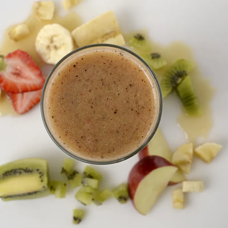 blended fruit in a glass, surrounded by slices of strawberry, banana, kiwi and red delicious apple