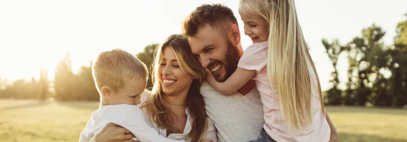 A family of four, the mother holding the son and the father holding the daughter, all laughing and smiling. The sun setting behind them.