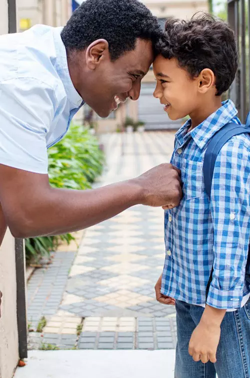 A father and a son standing outside,. The son is wearing a backpack and the father is bending over, comforting him.