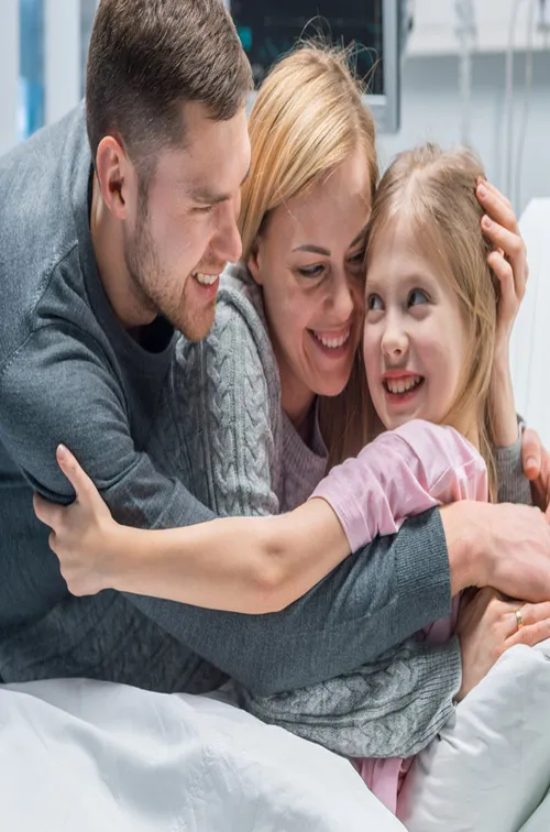 A family hugging their young daughter in her hospital bed.