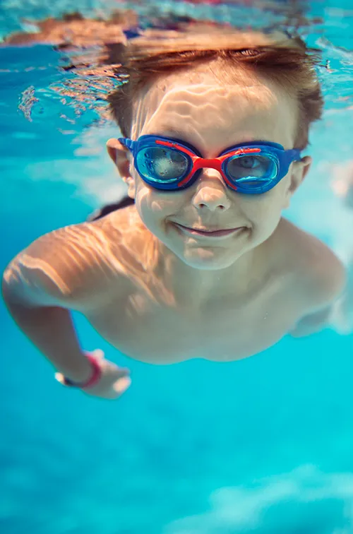 A young girl and boy swimming underwater in a pool while wearing goggles.