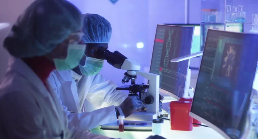 2 individuals in full ppe in a research lab face computers, one looks into a microscope