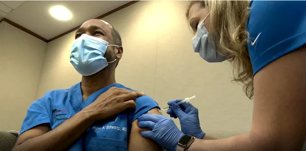 Dr. Schwartz rolls up his blue scrub sleeve as nurse administers the COVID-19 Vaccine